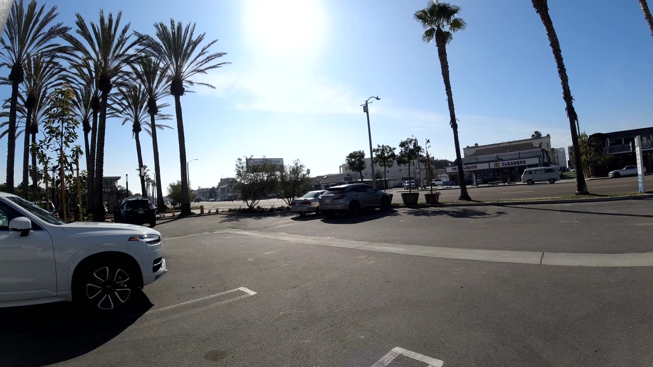 Huntington Beach frame with the white car more prominent at the right edge, a palm-lined road beyond it, several vehicles near storefronts, and no visible people.