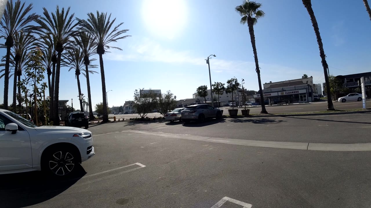 Huntington Beach street frame with the white foreground car slightly farther into view, cars along the opposite curb, palms, storefronts, and no visible people.