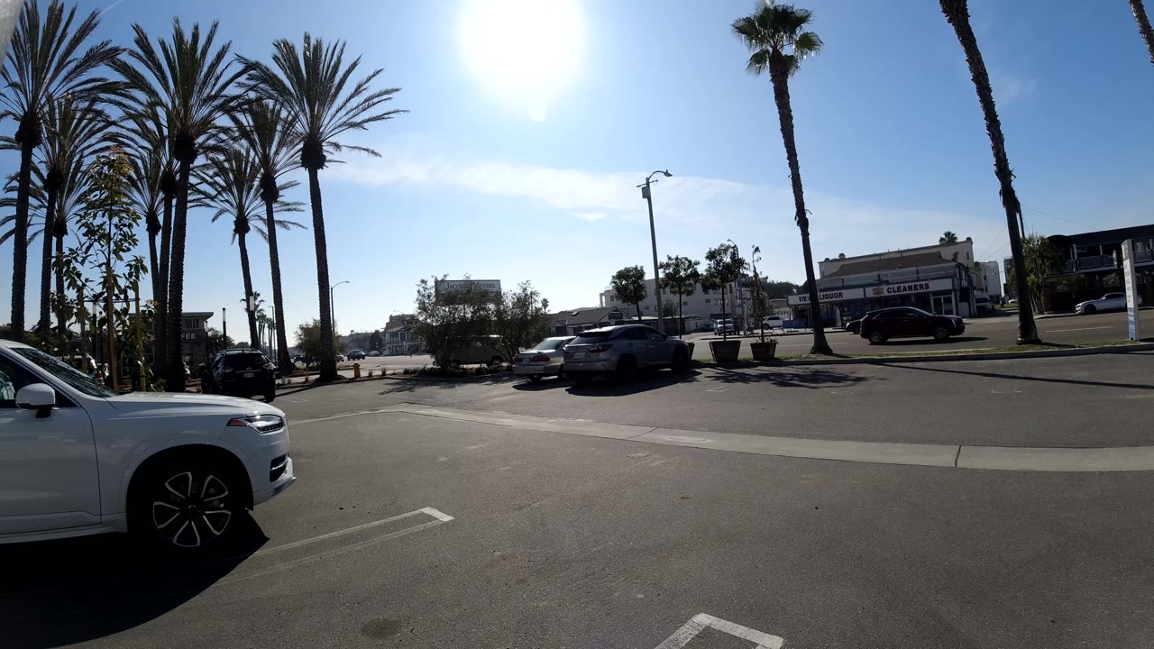 A wide Huntington Beach parking lot view shows palm trees, parked cars, a white car at the left edge, low buildings, bright blue sky, and no visible people.
