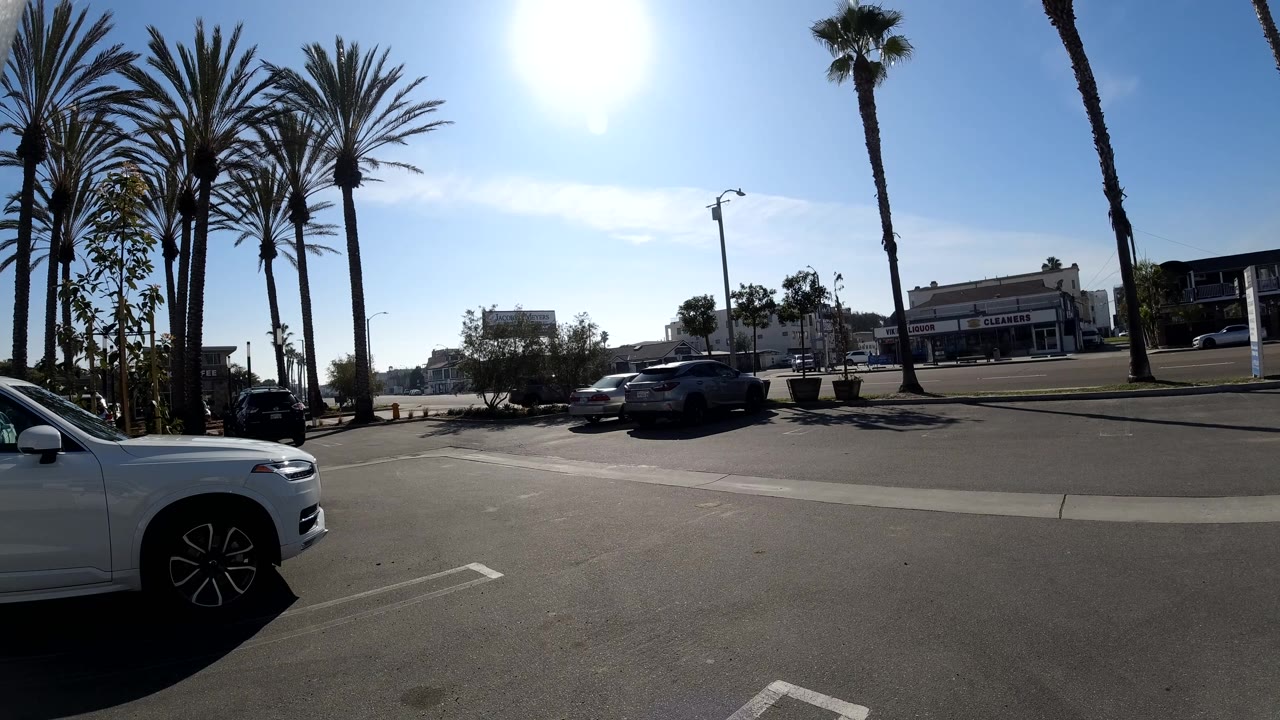 A bright Huntington Beach parking lot frame shows cars clustered near the street, palm trunks cutting through the sky, low storefronts in the distance, and no visible people.