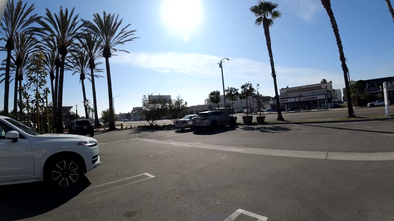 Second frame of the Huntington Beach street view, showing a white car cropped along the right edge, palms lining the road, vehicles in the midground, low buildings, bright blue sky, and no visible people.