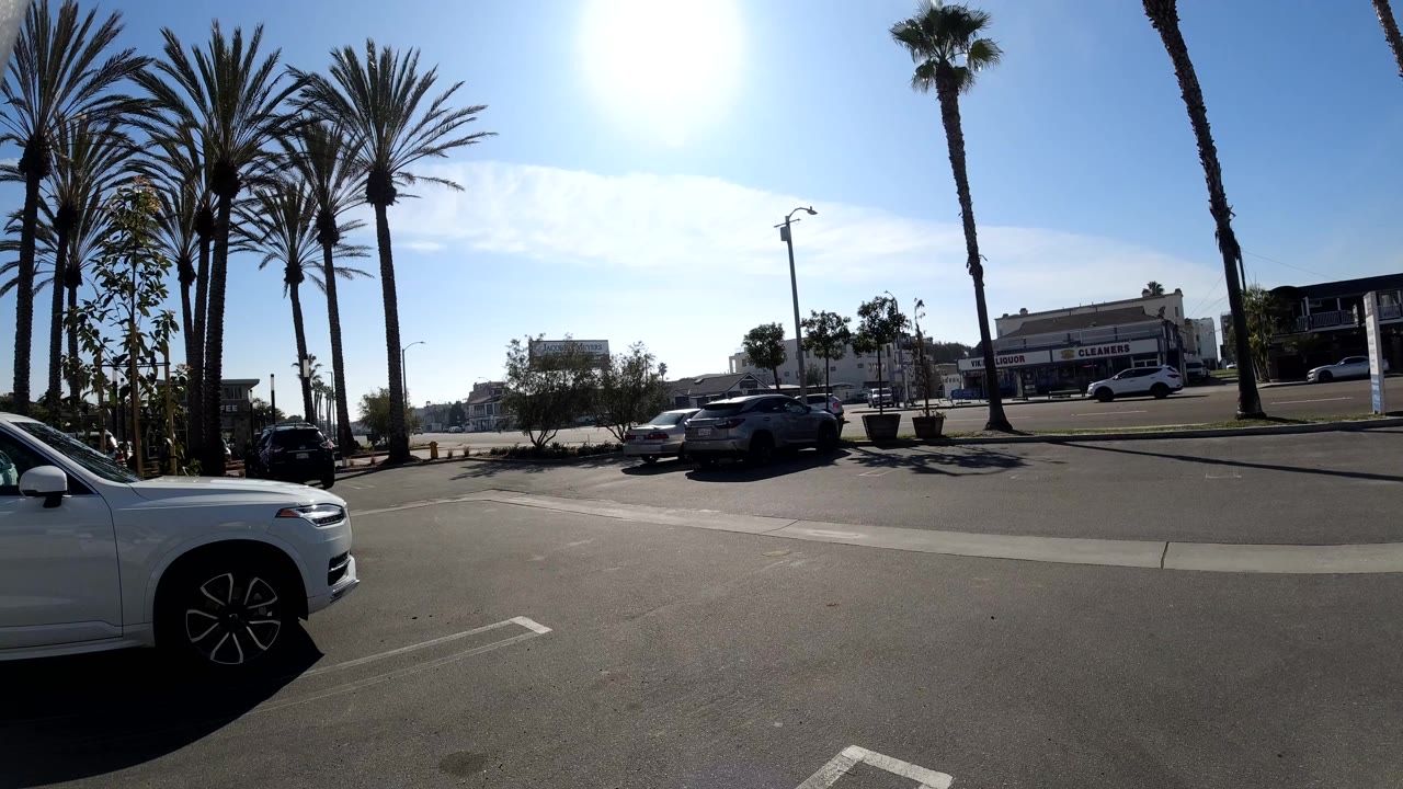 A wide Huntington Beach street view shows tall palm trees, a bright sky, parked cars, and the front of a white vehicle cropped at the lower left.