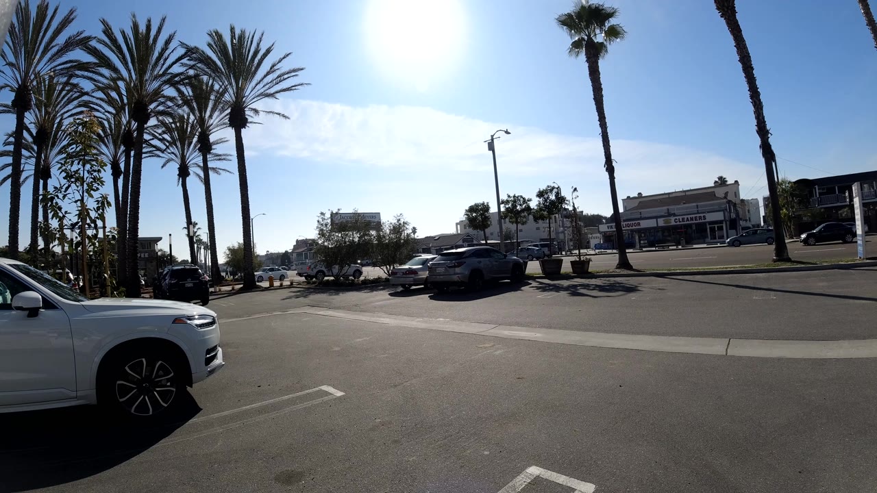 A wide Huntington Beach road frame shows moving and parked cars, palm trunks, a cropped white car at the lower right, and low buildings under hard sunlight.