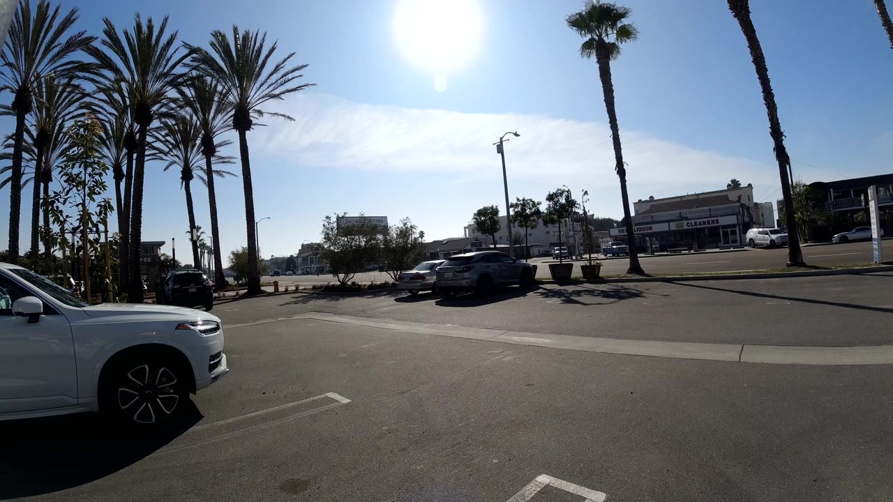 Parking lot scene with foreground white car, palms, parked vehicles, and storefronts.