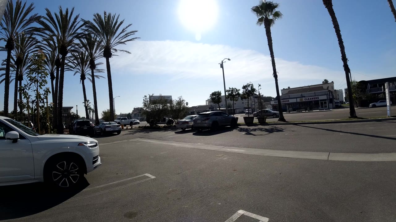 Opening frame of a Huntington Beach parking lot with a cropped white car at left, tall palms along the street, low retail shops in the background, and no visible people.