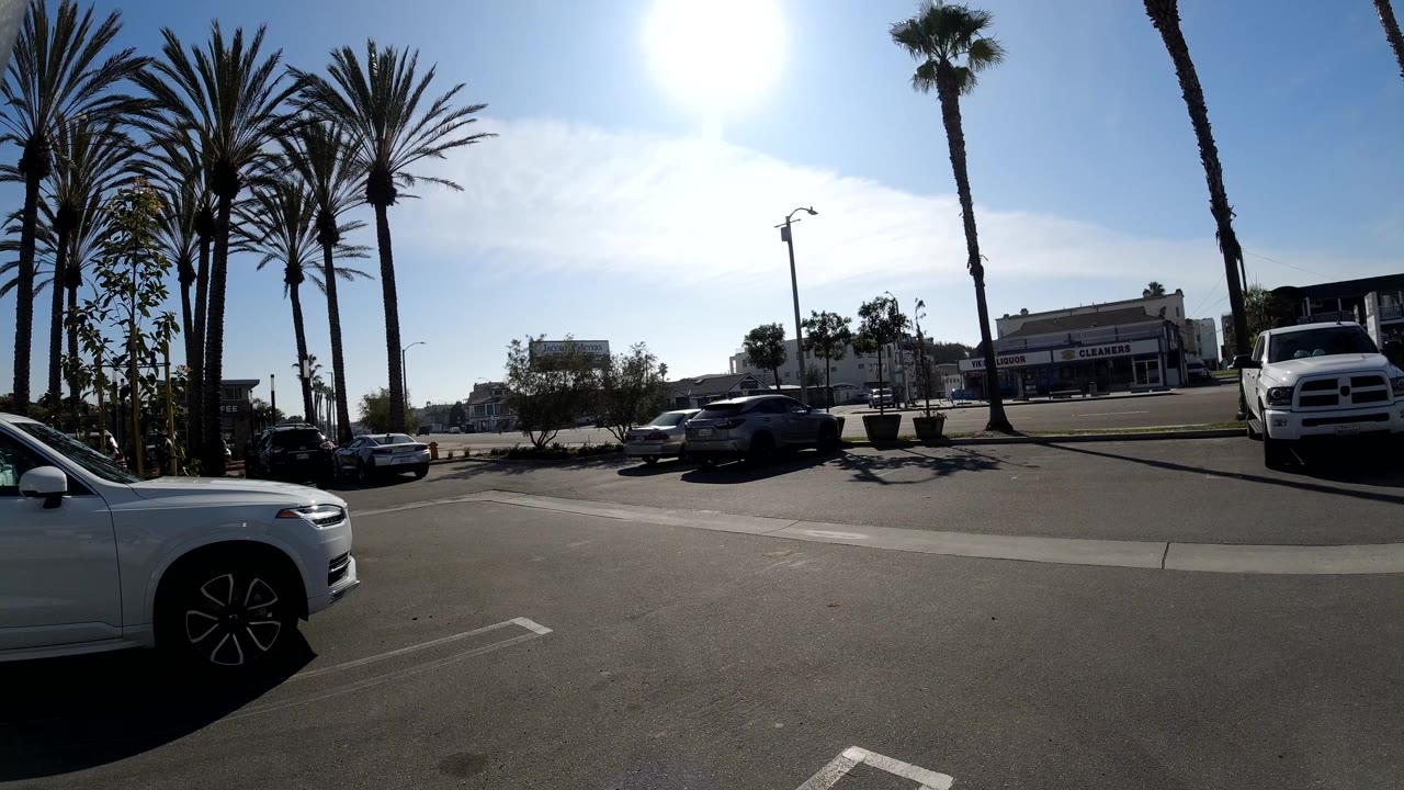 Start of a Huntington Beach street parking view with palm trees, parked cars, bright overhead sun, low buildings across the road, and no visible people.