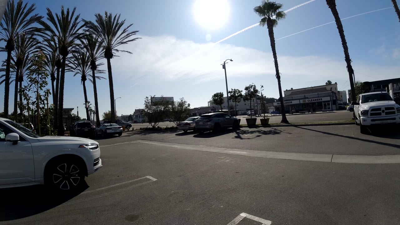 Opening frame of a sunny Huntington Beach road with tall palm trees, a white car at the left edge, parked vehicles, low commercial buildings, and no clearly visible people.