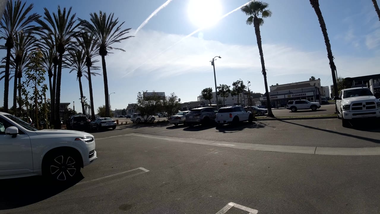 Second frame of the Huntington Beach parking lot view, with a white SUV cropped in the foreground, dark cars along the curb, pickup trucks to the right, palm trunks, and bright glare in the sky.