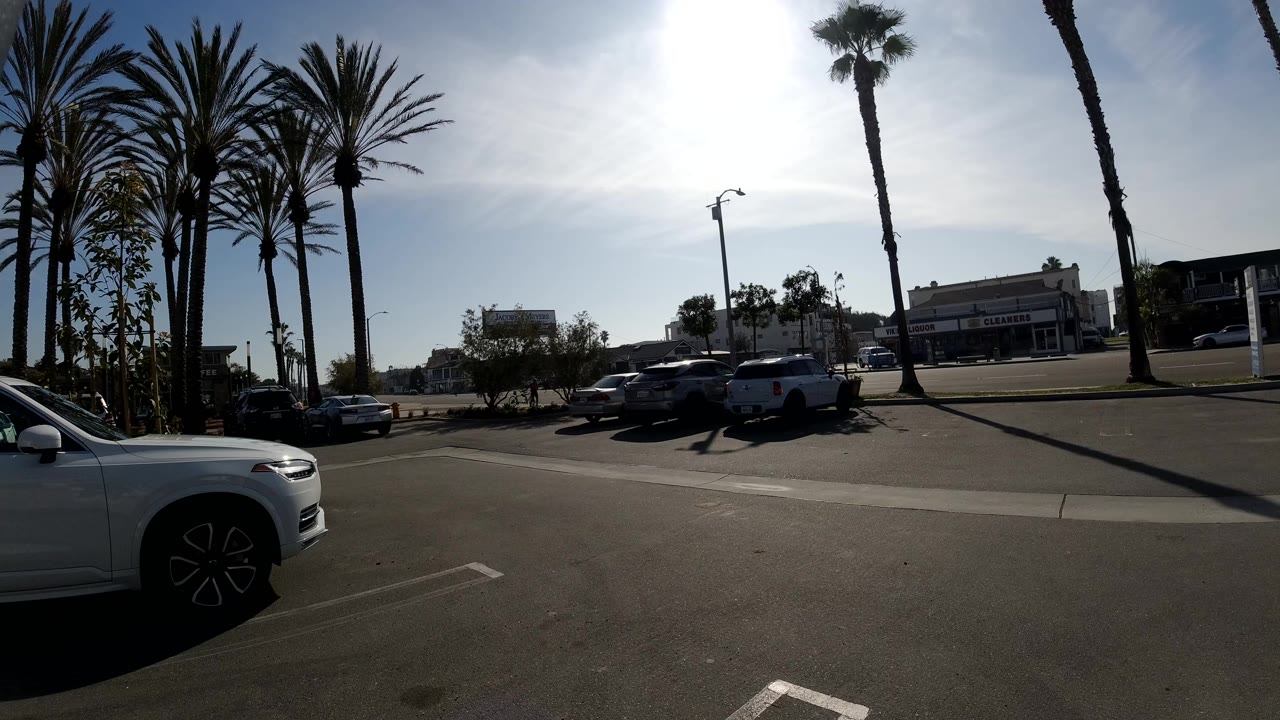 Huntington Beach parking lot view one second in, with tall palms silhouetted against glare, a white car at the lower edge, parked vehicles, and no visible people.