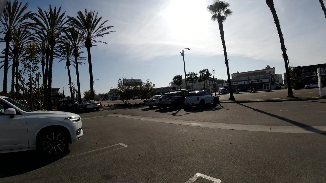 Opening frame of a Huntington Beach parking lot with tall palms at left, a white SUV cropped in the foreground, cars across the lot, low commercial buildings in the distance, and no visible people.