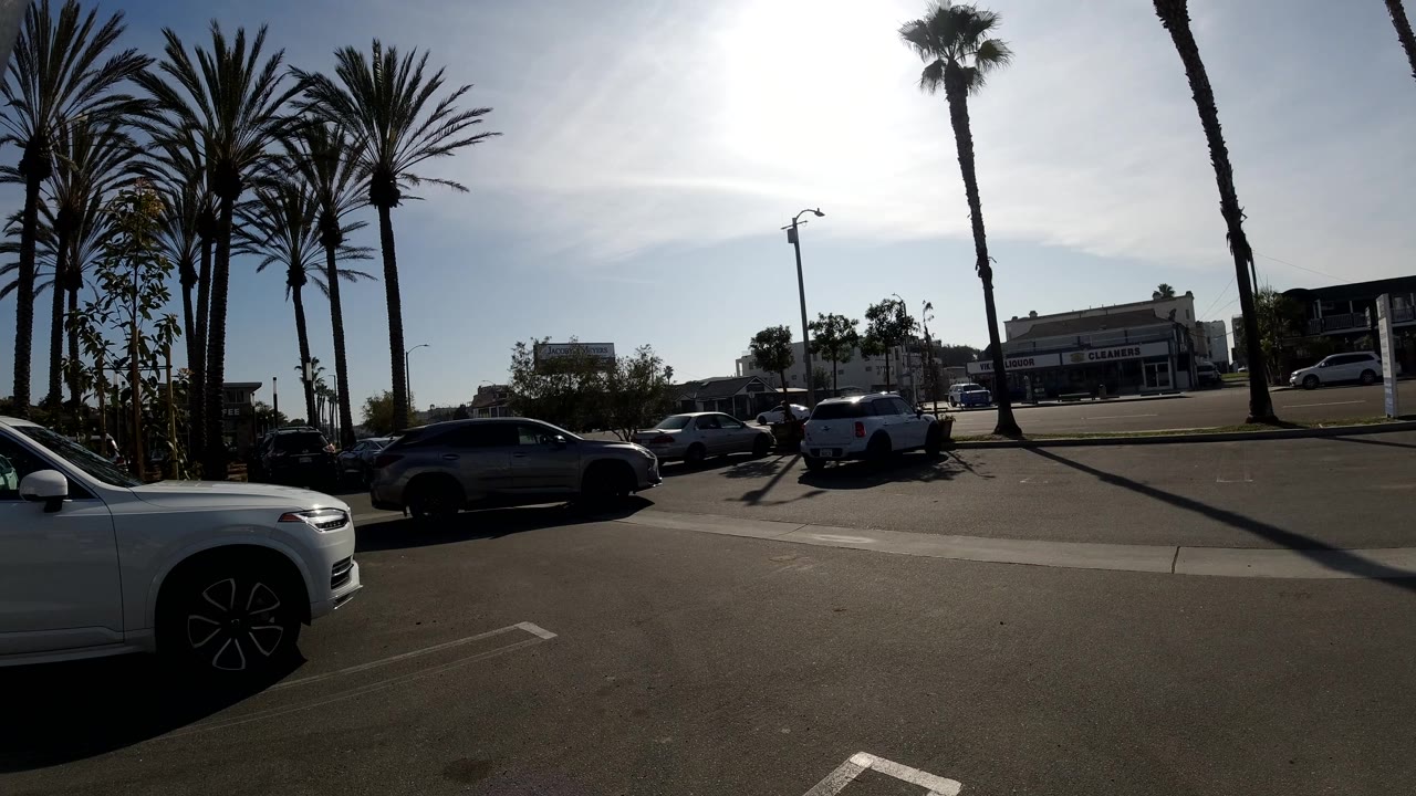 A dark vehicle moves through the right side of the Huntington Beach parking lot while the foreground white SUV, palm trees, and distant buildings remain in place, with no visible people.