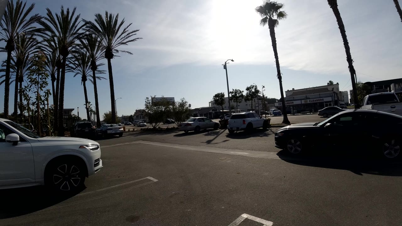 A Huntington Beach parking lot in bright sun shows a white SUV at the left edge, parked cars, tall palm trees, low buildings, and no visible people.