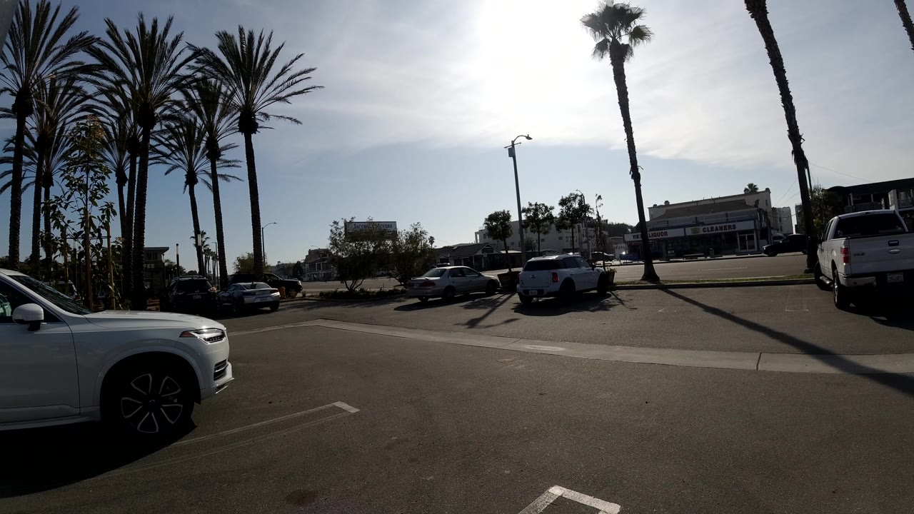 The passing car has cleared, leaving a Huntington Beach parking lot view with a white SUV at left, parked vehicles, tall palms, hard shadows, and no visible people.