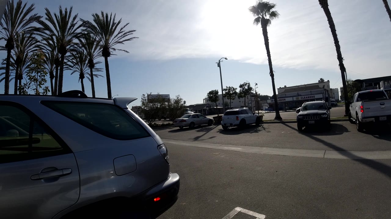 The dark pickup truck sits near the center of the Huntington Beach frame, with a foreground SUV, palm trunks, parked vehicles, low buildings, and high backlight shaping the view.