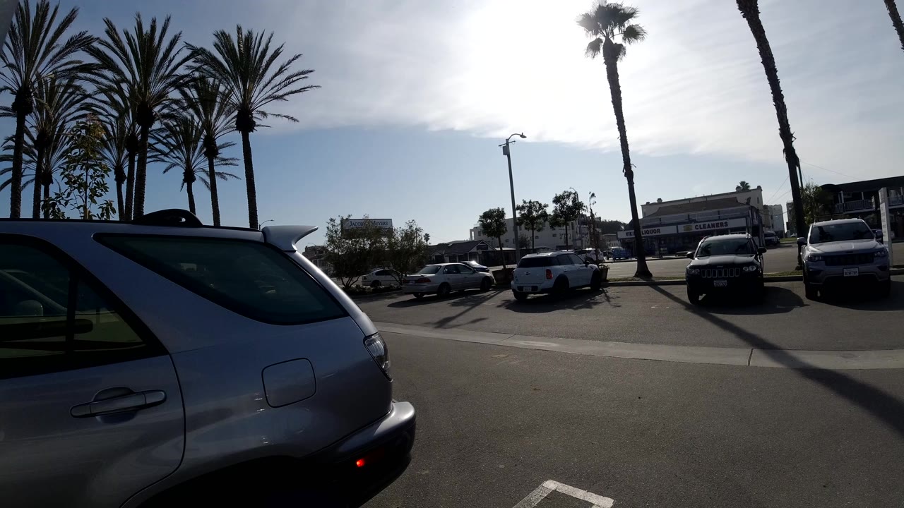 A silver SUV fills the left foreground of a Huntington Beach parking lot, with palm trees, parked vehicles, bright sky, and no visible people.