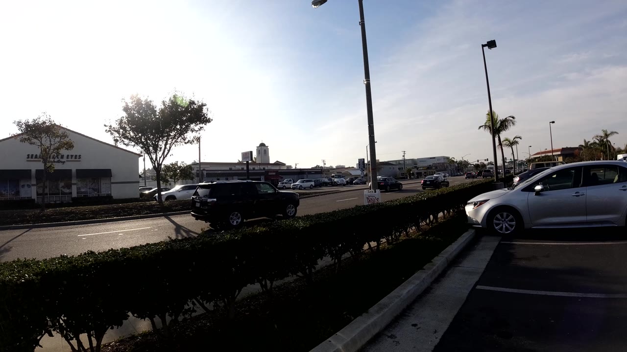 Bright Bolsa Chica parking area with strong sun glare, a hedge in the foreground, cars along the road, street lights, low commercial buildings, and no visible people.