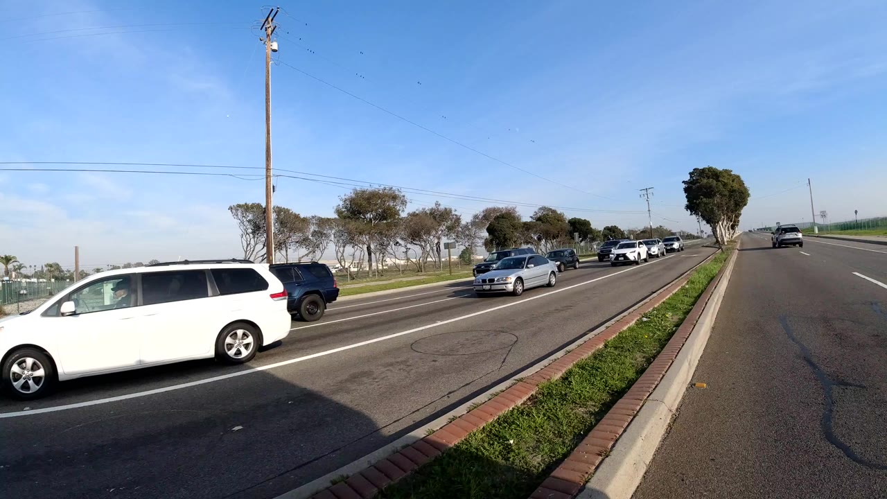 Cars travel along a divided Bolsa Chica, California roadway beside a grassy median, trees, utility poles, and clear blue sky, with no visible people.