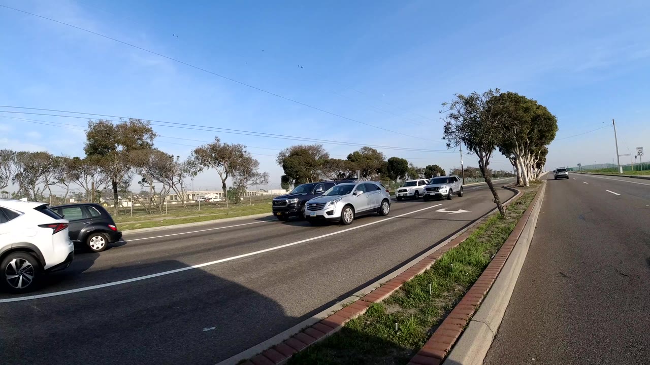 A white car and other vehicles pass on a broad Bolsa Chica, California road with a low median, small trees, utility lines, and no visible people.