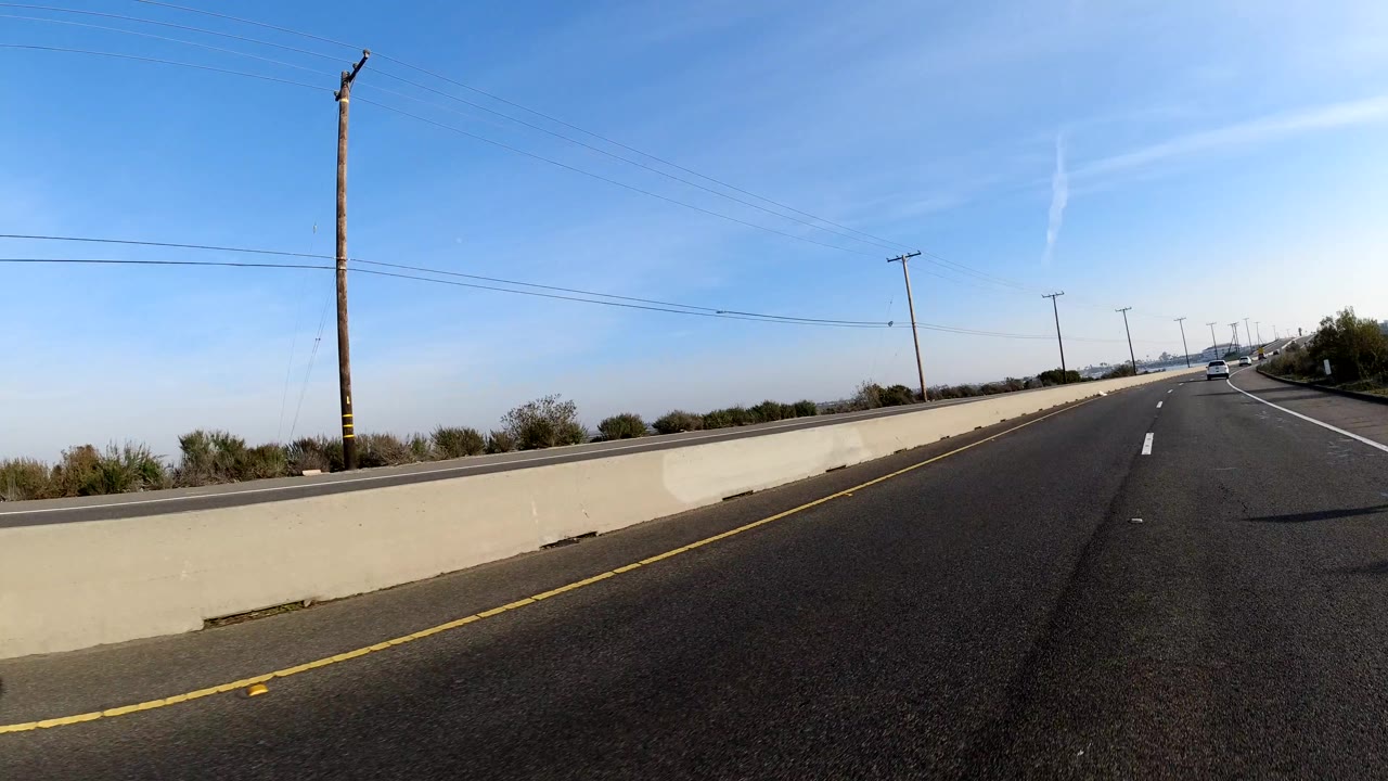 The view shifts to an open Bolsa Chica, California roadway bordered by a concrete barrier, utility poles, lane markings, distant cars, and no visible people.