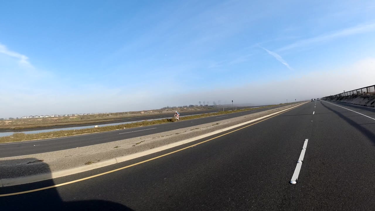 Open divided road beside flat wetlands and a distant bridge.