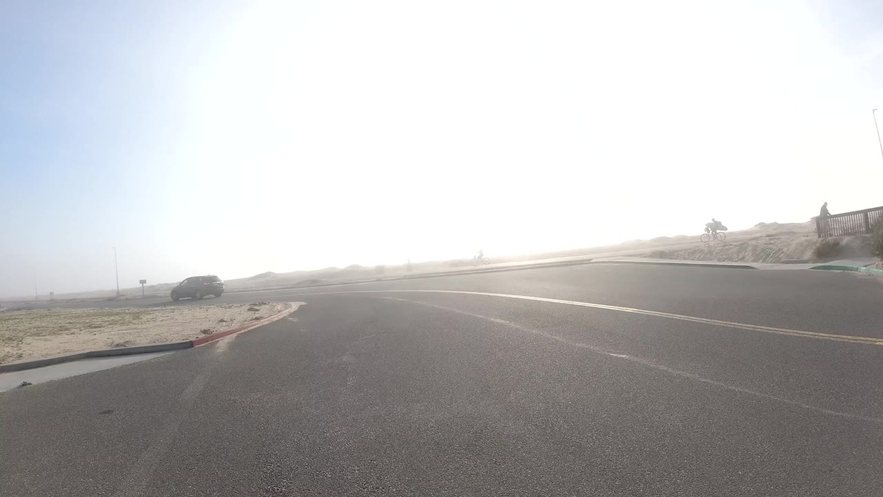 A wide bright road scene in Bolsa Chica, California shows open pavement, strong glare, a distant vehicle, sandy roadside, and no visible people.