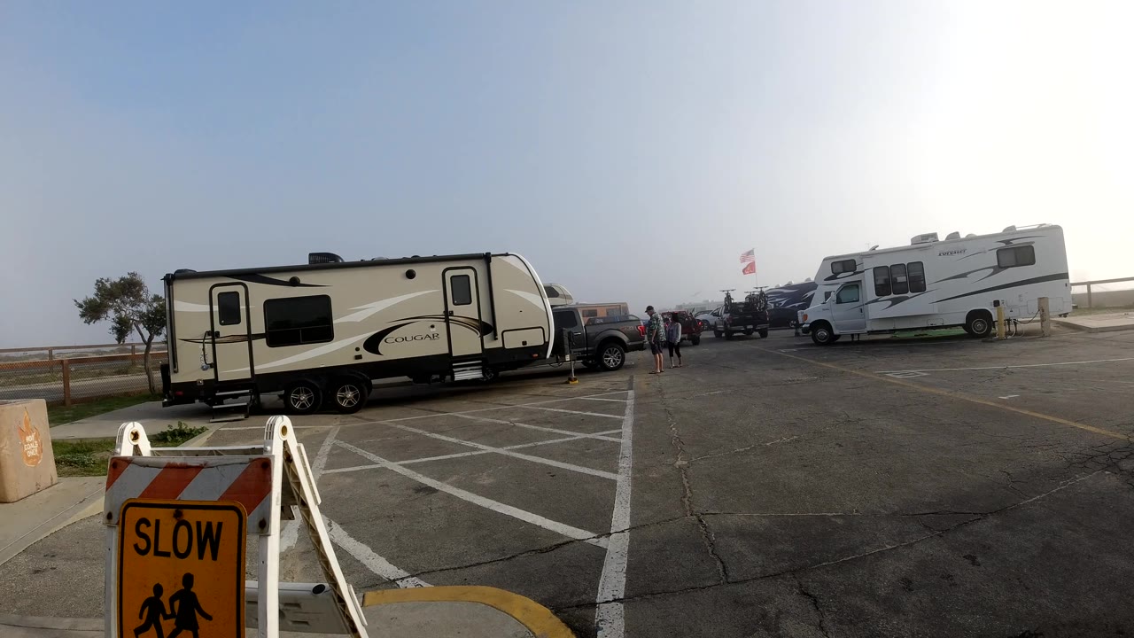 Wide Bolsa Chica, California lot with a travel trailer at left, RVs and parked cars across the midground, open pavement, hazy sky, and tiny distant figures near the vehicles.