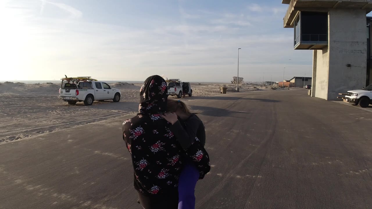 At 0:01 in Bolsa Chica, California, the woman and child remain centered in the foreground as white trucks sit near sandy ground and a concrete tower frames the right side.