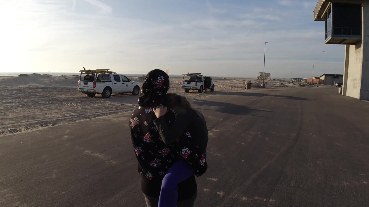 At 0:02 in Bolsa Chica, California, the woman carrying the child appears slightly farther right, with two white trucks on the sandy shoulder and the concrete tower at the edge of frame.
