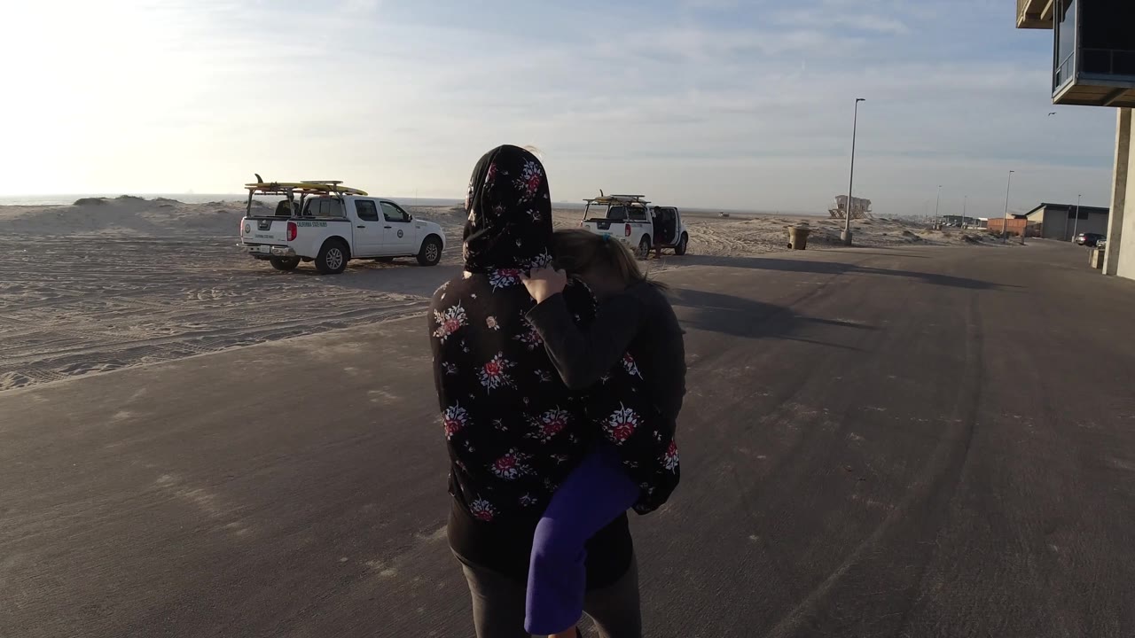 Woman carrying a child on a road with white trucks and open sand behind her.