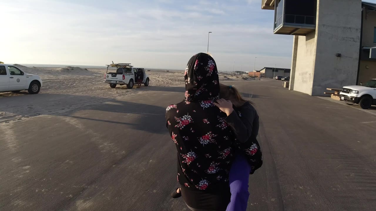 The woman and child remain centered from behind on a paved road in Bolsa Chica, California, with a white pickup at left, another truck near the sand, and the building edge at right.