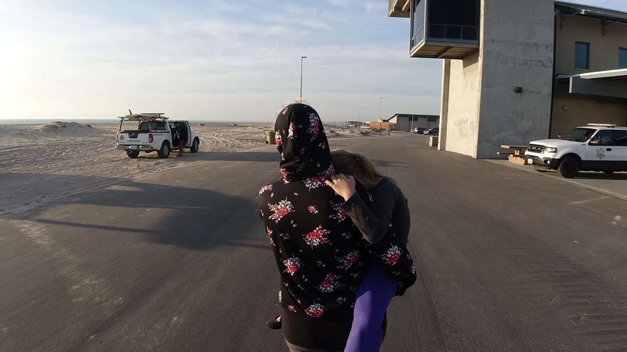 A rear view in Bolsa Chica, California shows the woman carrying the child closer to the right side of the road, with sandy ground and parked white pickup trucks spread across the background.
