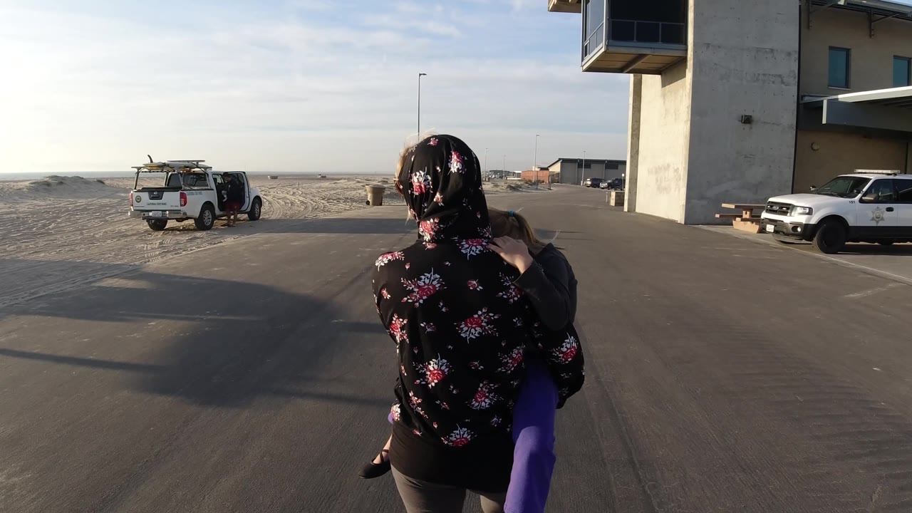 Woman in a floral head covering carries a child past parked trucks near sand.