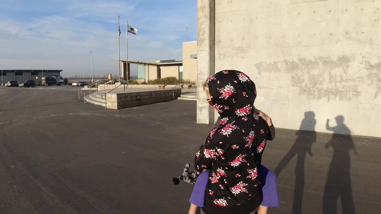At 0:00 in Bolsa Chica, California, a mother in a black floral hood carries a young child on her shoulders near a concrete wall, with flagpoles and parked vehicles across the lot.