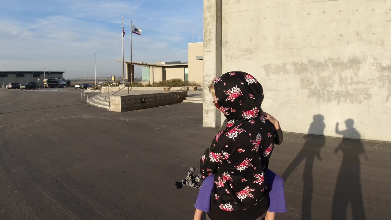 At 0:01 in Bolsa Chica, California, the mother and child are seen from behind beside the concrete wall, with two long shadows cast on the pavement and a low building beyond the parking area.