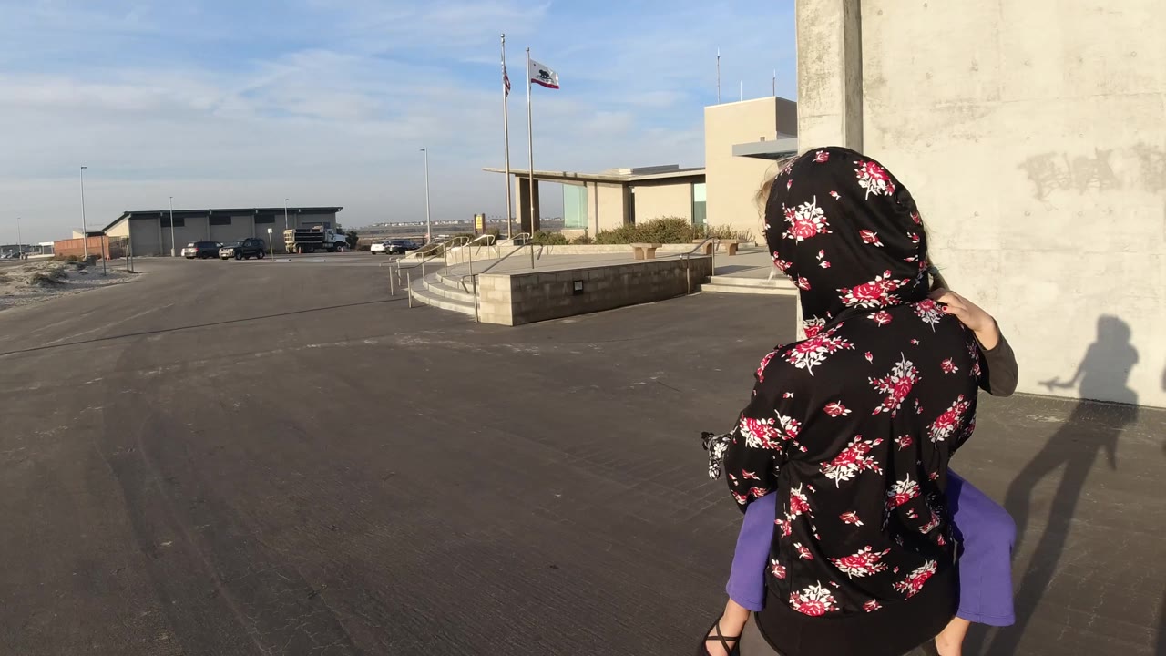 At 0:02 in Bolsa Chica, California, the child rests on the mother's shoulders as they stand near the wall, with sunlit pavement, flagpoles, parked vehicles, and the building entrance area visible.