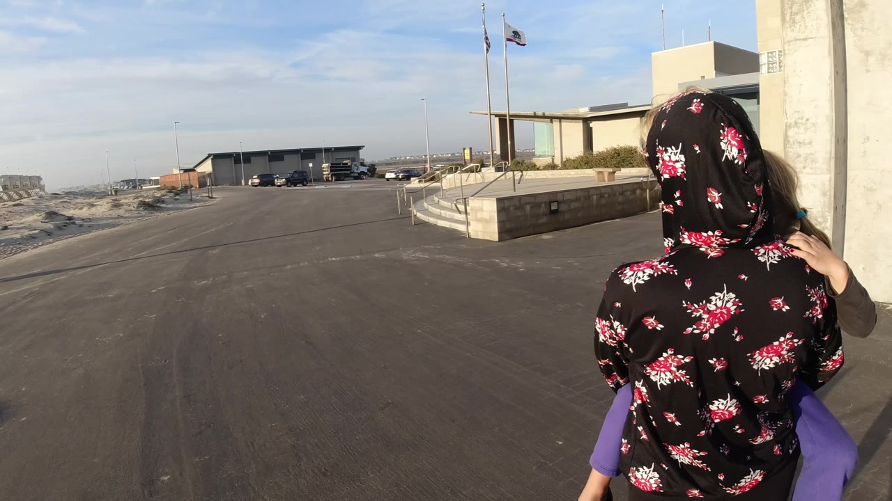 Mother carrying a child on her shoulders near a building entrance and open lot.