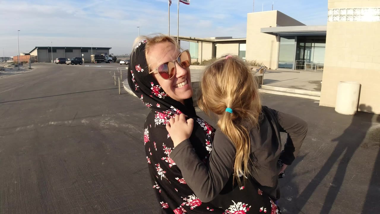 In Bolsa Chica, California, a smiling woman in sunglasses carries a young blond girl beside a beige public building and paved roadway.
