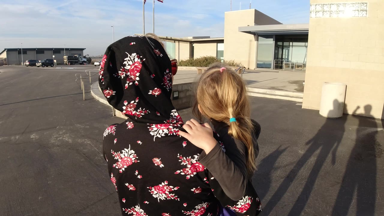 In Bolsa Chica, California, the camera follows behind the woman and child as they face the building, with their long shadows falling across the pavement.