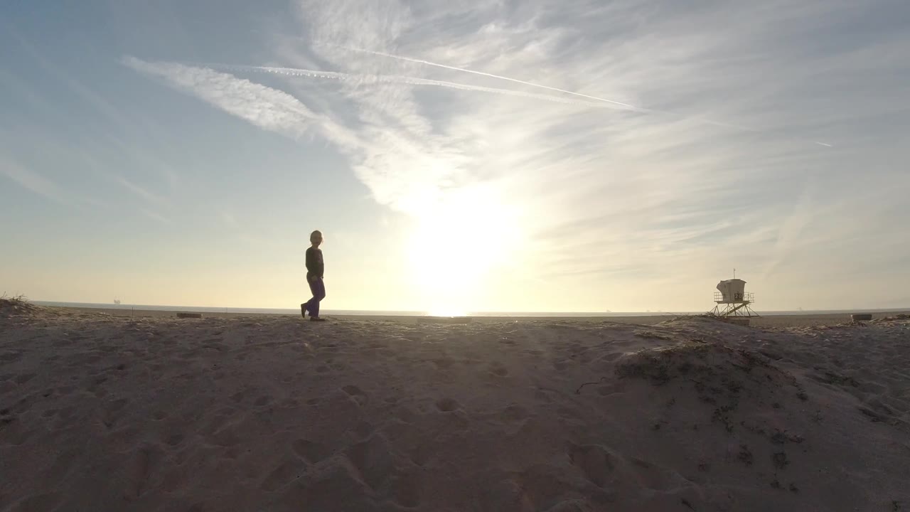 At Bolsa Chica, California, a young girl stands on open sand with a bright low sun behind thin clouds and a distant beach tower to the right.
