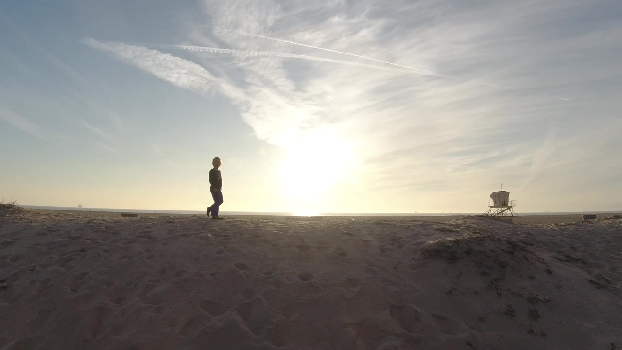 At Bolsa Chica, California, the girl remains on the open beach as the bright sun flares near the horizon and a small tower sits in the far right background.