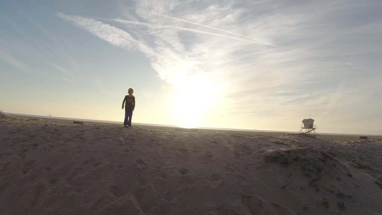 At Bolsa Chica, California, the young girl faces nearer toward the camera on the sandy beach while the low sun glows behind her and the distant tower remains to the right.