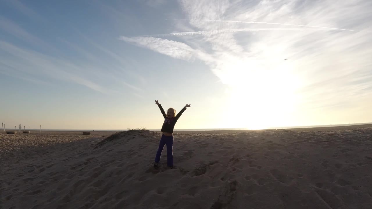 A person stands on sandy terrain at Bolsa Chica, California with arms lifted under a bright low sun and streaked clouds at the start of the clip.