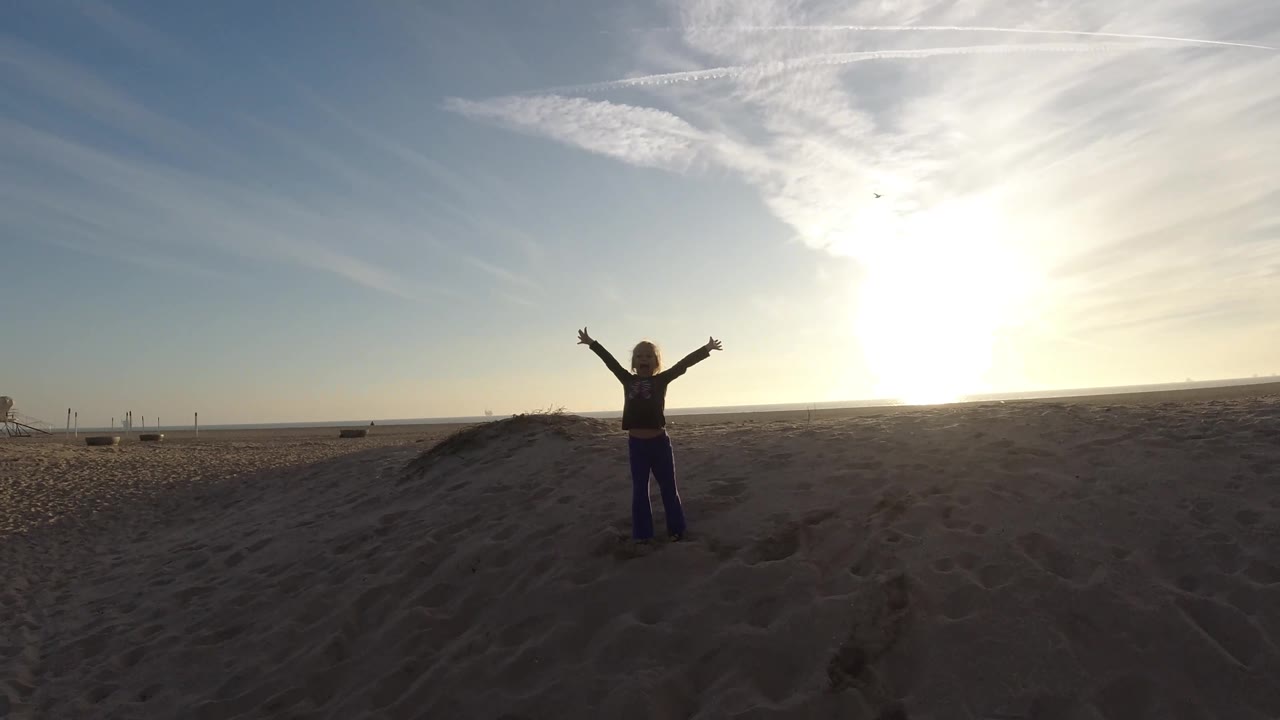 The person appears farther right in the wide Bolsa Chica beach view, with the low sun flaring near the horizon and open sand stretching across the foreground.