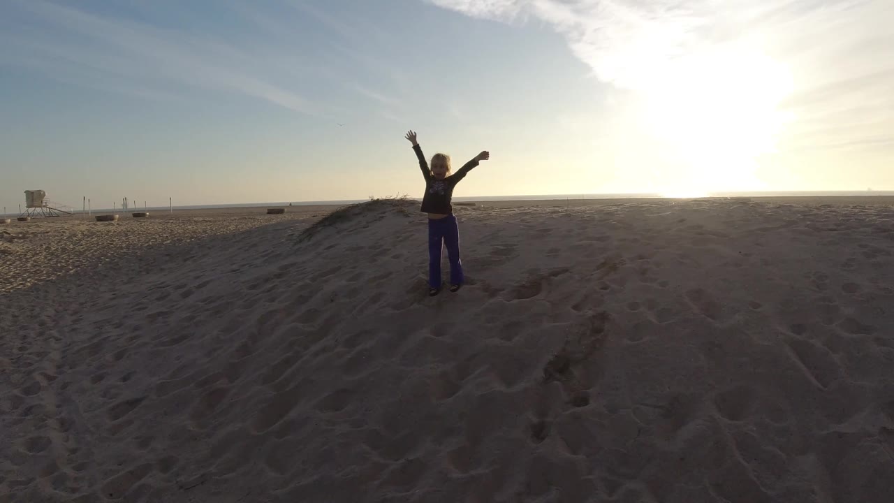 At Bolsa Chica, California, a single person stands on open sand with one arm high and one arm out while the sun flares from the upper right.