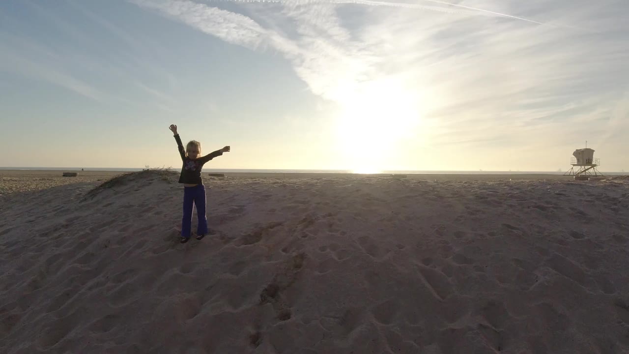 A young girl stands on Bolsa Chica sand with one arm raised and one arm extended as the low sun backlights the beach and a lifeguard tower sits on the horizon.