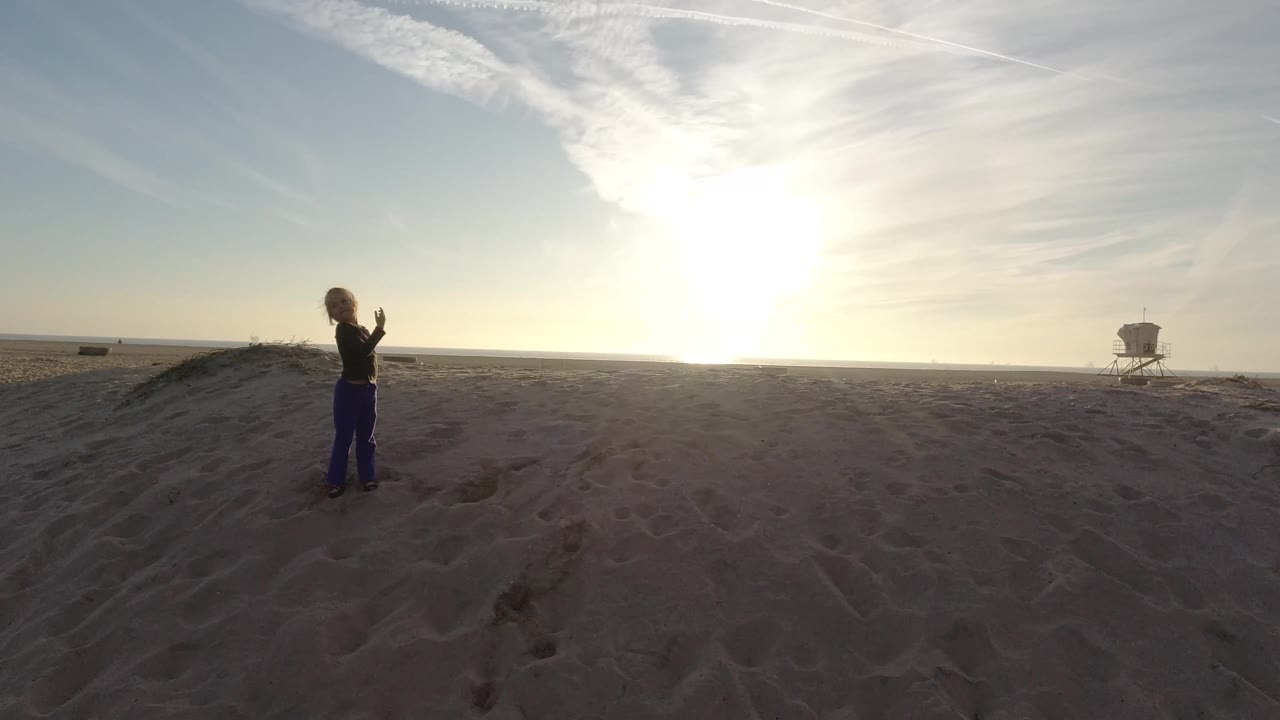 The child has turned sideways on the Bolsa Chica sand, with hands near her chest, a small sand mound to the right, and the lifeguard tower farther along the horizon.