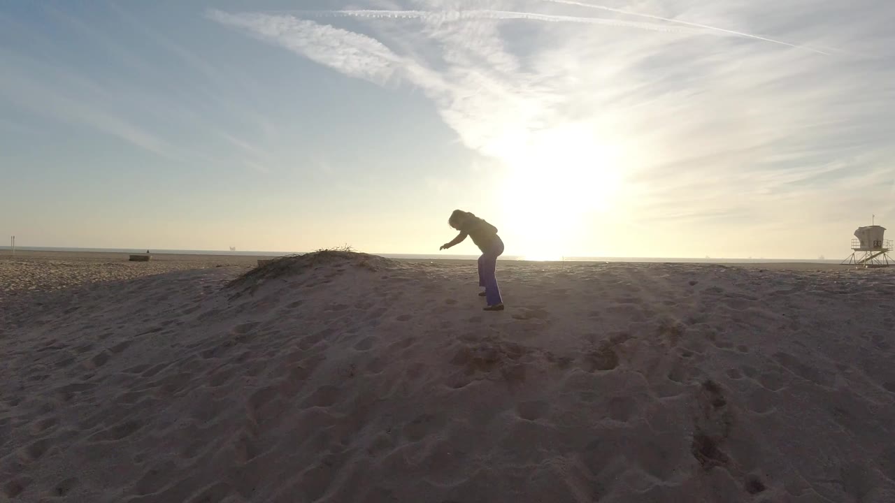 Backlit child on a sand mound with sunset glare and a distant lifeguard tower.