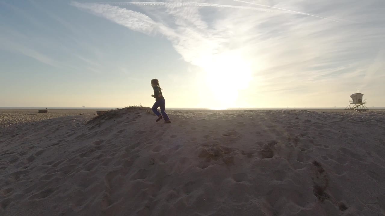 A child runs across the Bolsa Chica sand in silhouette while the low sun washes the sky and a lifeguard tower stands near the right side of the frame.