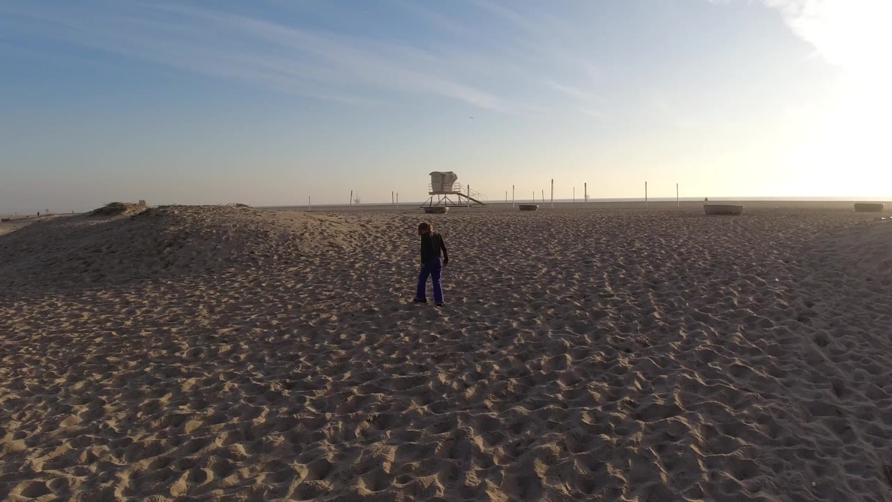 At Bolsa Chica, California, an adult and child appear small in a wide beach frame, with rippled sand in front and a lifeguard tower near the horizon.