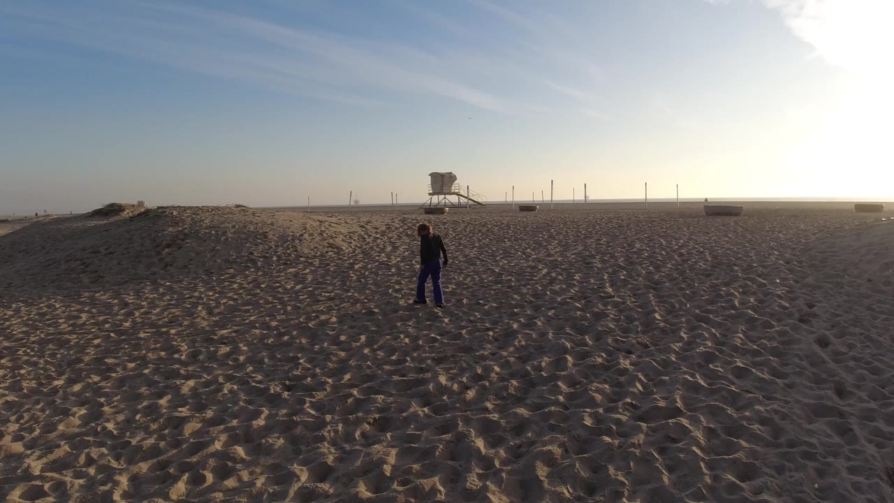The adult and child remain near the center of the Bolsa Chica beach view, with patterned sand leading back to the lifeguard tower and bright haze at right.